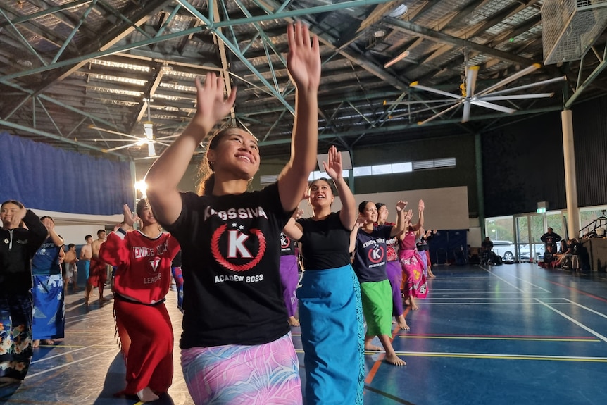 Rows of young women practice Siva Samoa in school hall. Close up to camera is young woman wearing blue tee and ie lava lava.