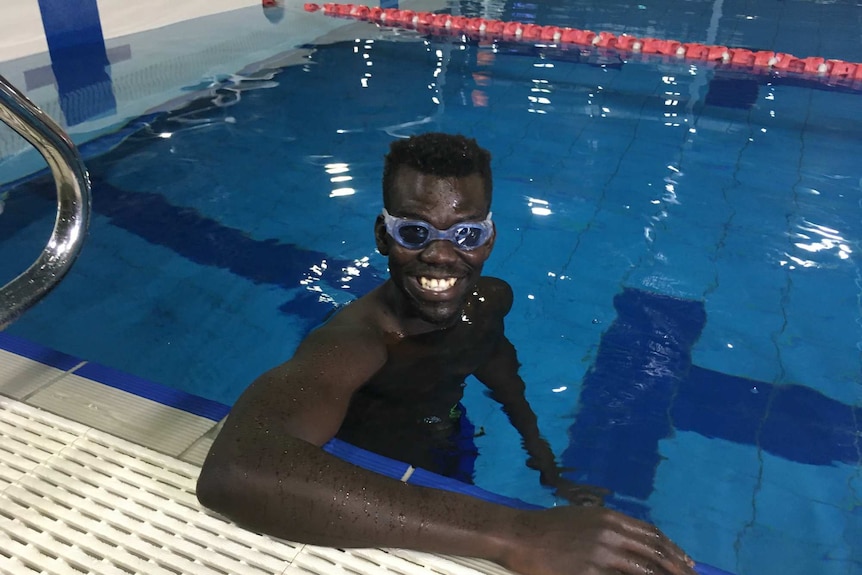 Mella Opiew, wearing swimming goggles, stands in the water with his hand on the side of the pool, smiling at the camera.