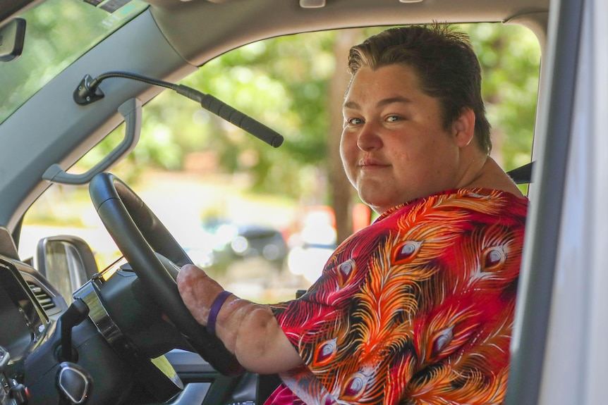 A woman with fair skin and short hair wearing red, who has had her legs and hands amputated, sits in the drivers seat of a car.