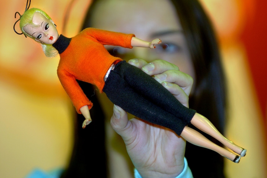 A close up of a doll with blonde hair wearing an orange jumper and black pants.