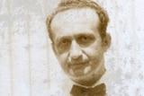 Melekh Ravitsh, wearing a white shirt, bow tie, stands in front of a corrugated iron wall.