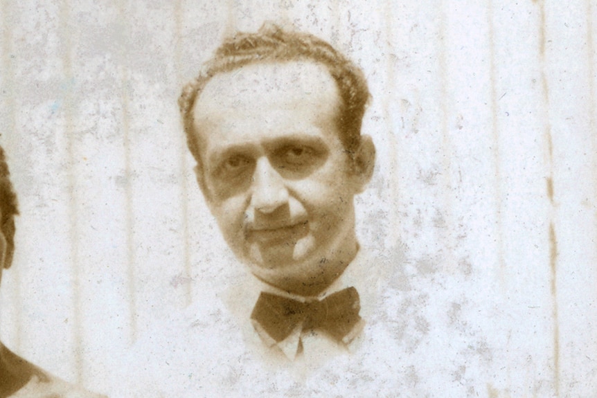 Melekh Ravitsh, wearing a white shirt, bow tie, stands in front of a corrugated iron wall.