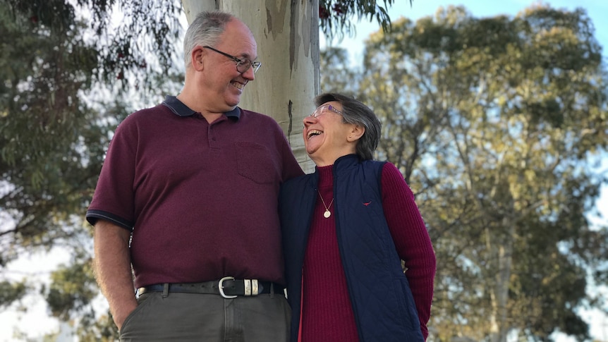 A man and a woman wearing burgundy smile at each other in front of a tree with a blue sky in the background.