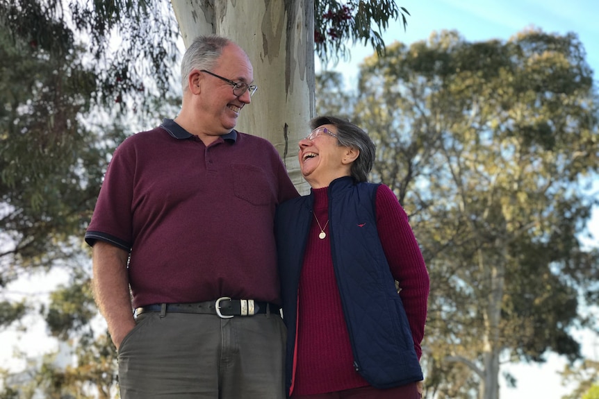 A man and a woman wearing burgundy smile at each other in front of a tree with a blue sky in the background.