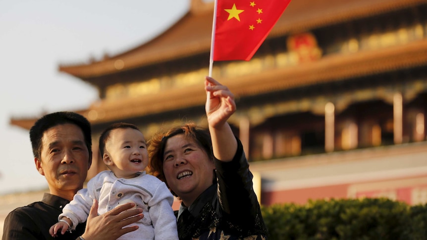 A child and his grandparents take a souvenir picture in front of the Tiananmen Gate. The grandmother waves a Chinese flag.