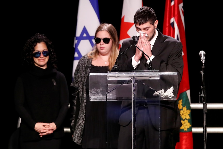 A man blows his nose standing at a lectern while a woman in sunglasses stands behind him