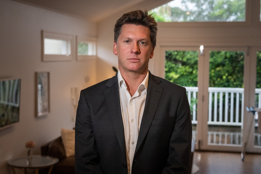 Gareth Aird stands in his house, wearing a suit but no tie. Green leaves are visible through a window behind him