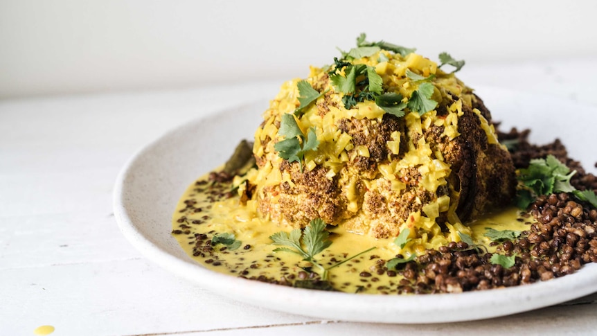 A plate with a whole roasted head of cauliflower, covered in tumeric sauce, coriander leaves, lentils, a vegetarian main.