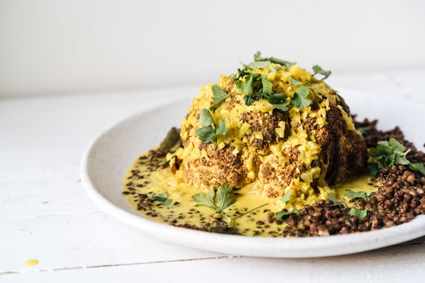 A plate with a whole roasted head of cauliflower, covered in tumeric sauce, coriander leaves, lentils, a vegetarian main.