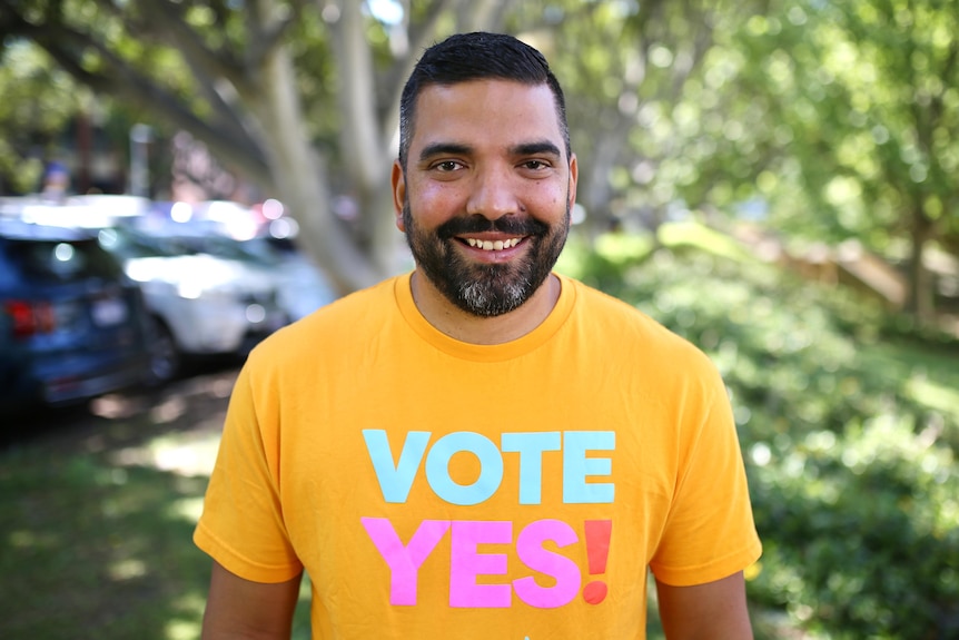 A tanned man with a yellow 'Vote Yes!' shirt smiles at the camera, with greenery in the background.