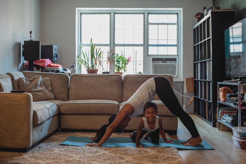 A young woman is in a yoga pose in a loungeroom with a small girl playing under her.