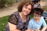 Sonia Sofianopoulos smiling while sitting on grass with her grandson Adam who is eating a sandwich.