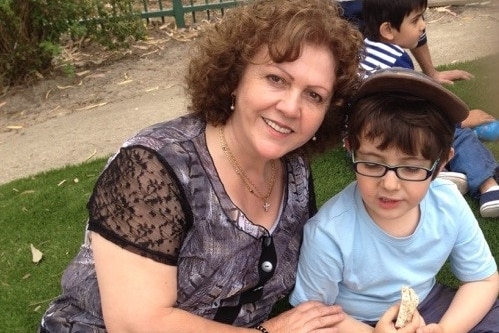 Sonia Sofianopoulos smiling while sitting on grass with her grandson Adam who is eating a sandwich.