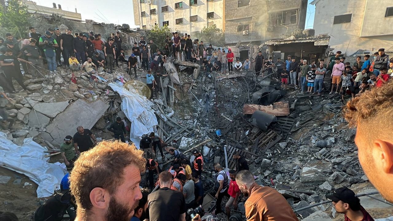 A large crowd of people gather around a house-sized hole in the ground filled with rubble and wreckage.