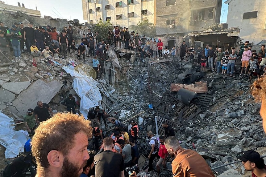 A large crowd of people gather around a house-sized hole in the ground filled with rubble and wreckage.