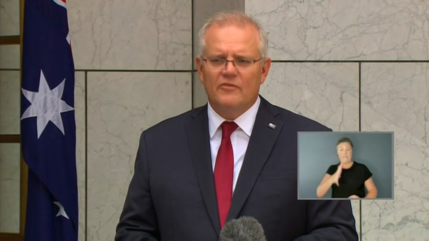Scott Morrison wearing a red tie stands beside an Australia flag.