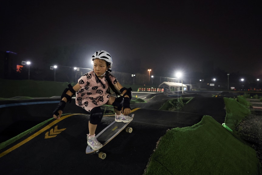 A young girl rides a skateboard at night on a pump track. 