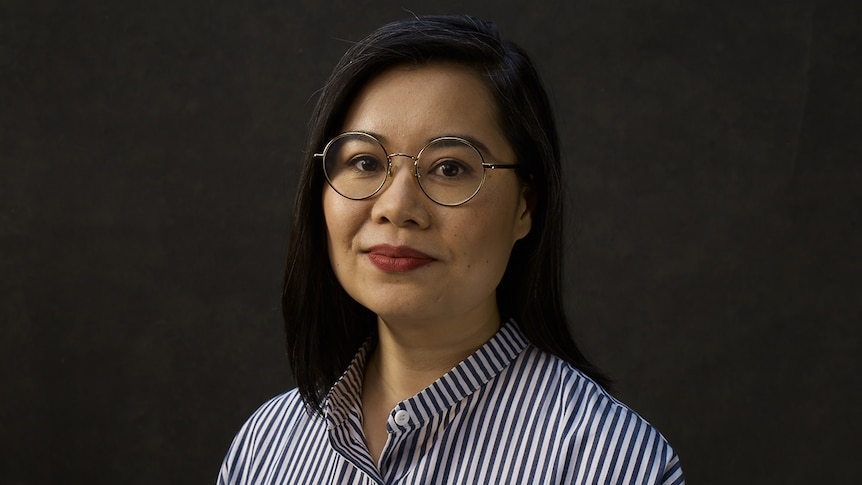 A woman wearing round metal glasses and white and blue striped shirts stands in front of dark grey wall.