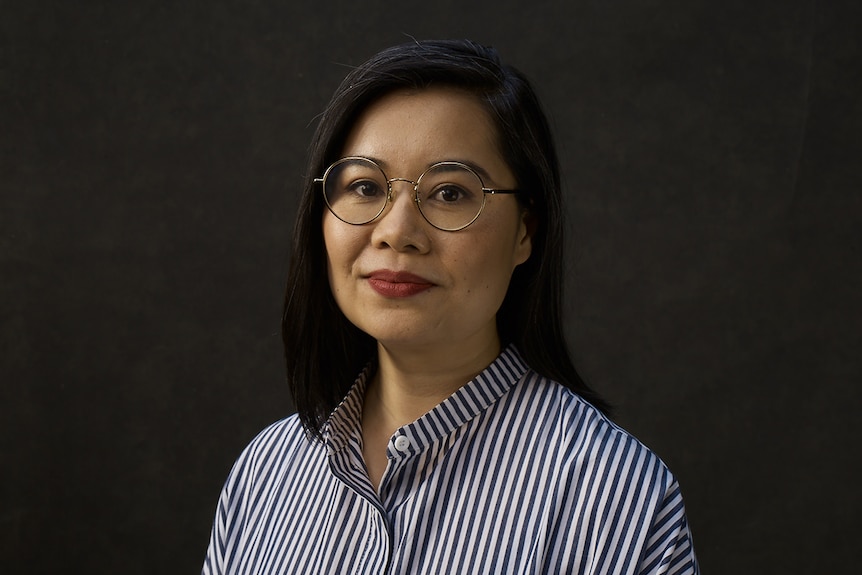 A woman wearing round metal glasses and white and blue striped shirts stands in front of dark grey wall.