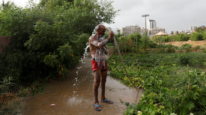 A slim South Asian man wearing red shorts cools off with hose over his head next to green park with city buildings behind