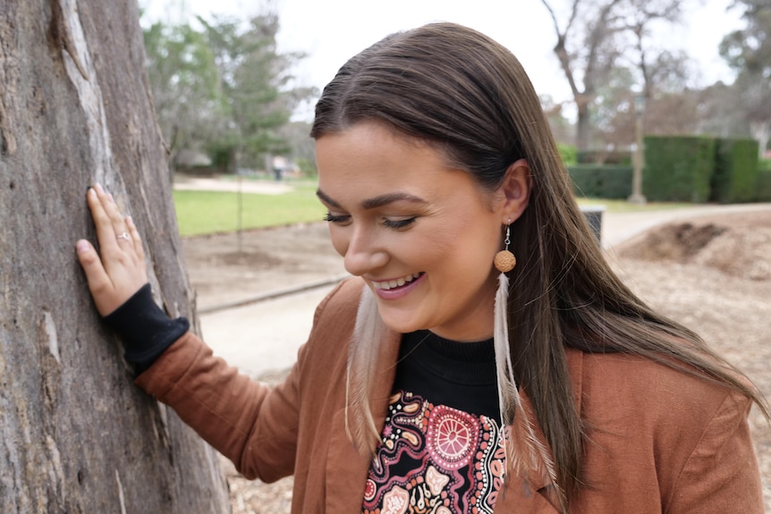 A woman wearing quandong and emu feather earrings leans against a gum tree with her head down and smiles. 