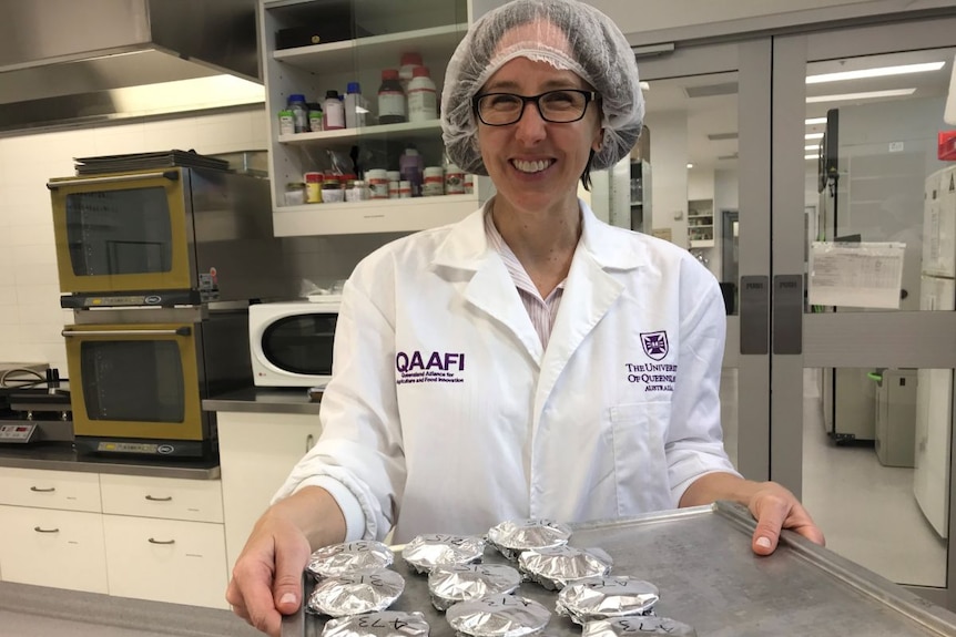 A smiling woman in a white lab coat and hair net holds a tray of tin-foil wrapped samples.