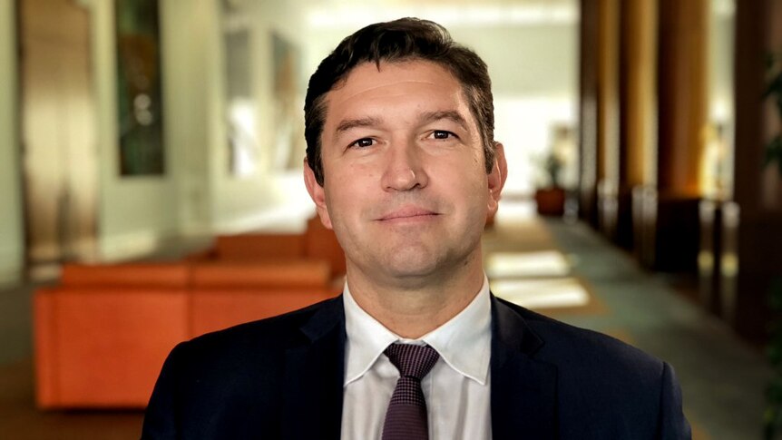 Man in suit and tie with black hair sits smiling with couches and formal walkway behind him