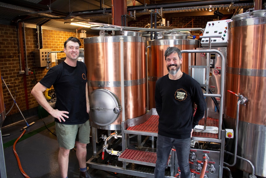 Two men in black shirts standing and smiling in front of brewery tanks.