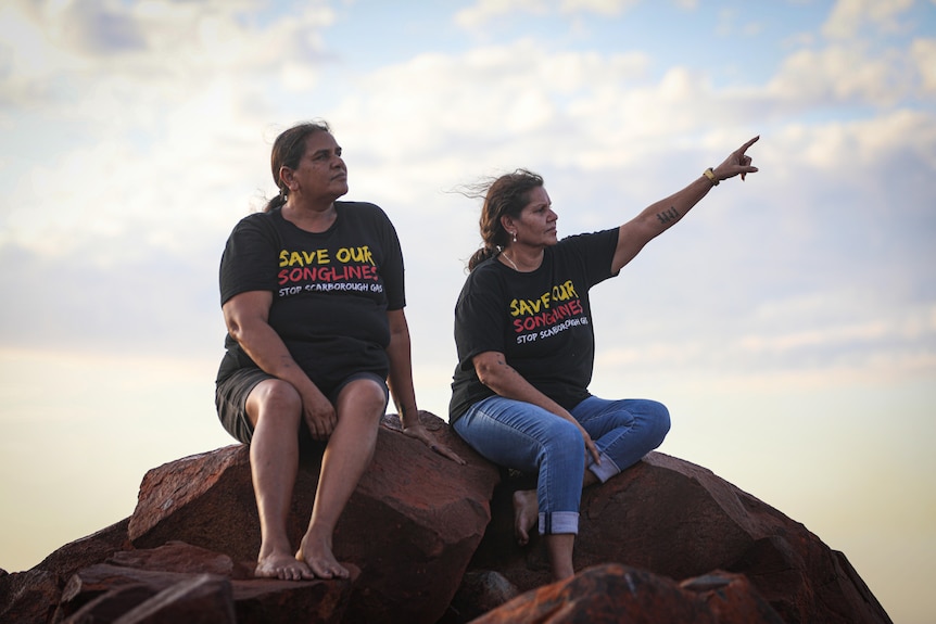 Two women sit on a large rock looking off into the distance, as one extends her arm and points.
