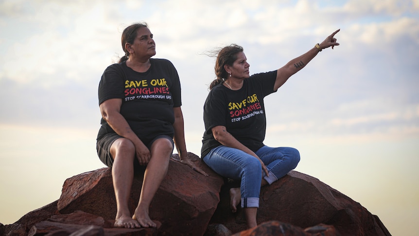 Two women sit on a large rock looking off into the distance, as one extends her arm and points.