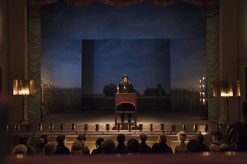 A man in Victorian era suit and ascot stands at podium in from of set and seated crowd in Regency era playhouse.