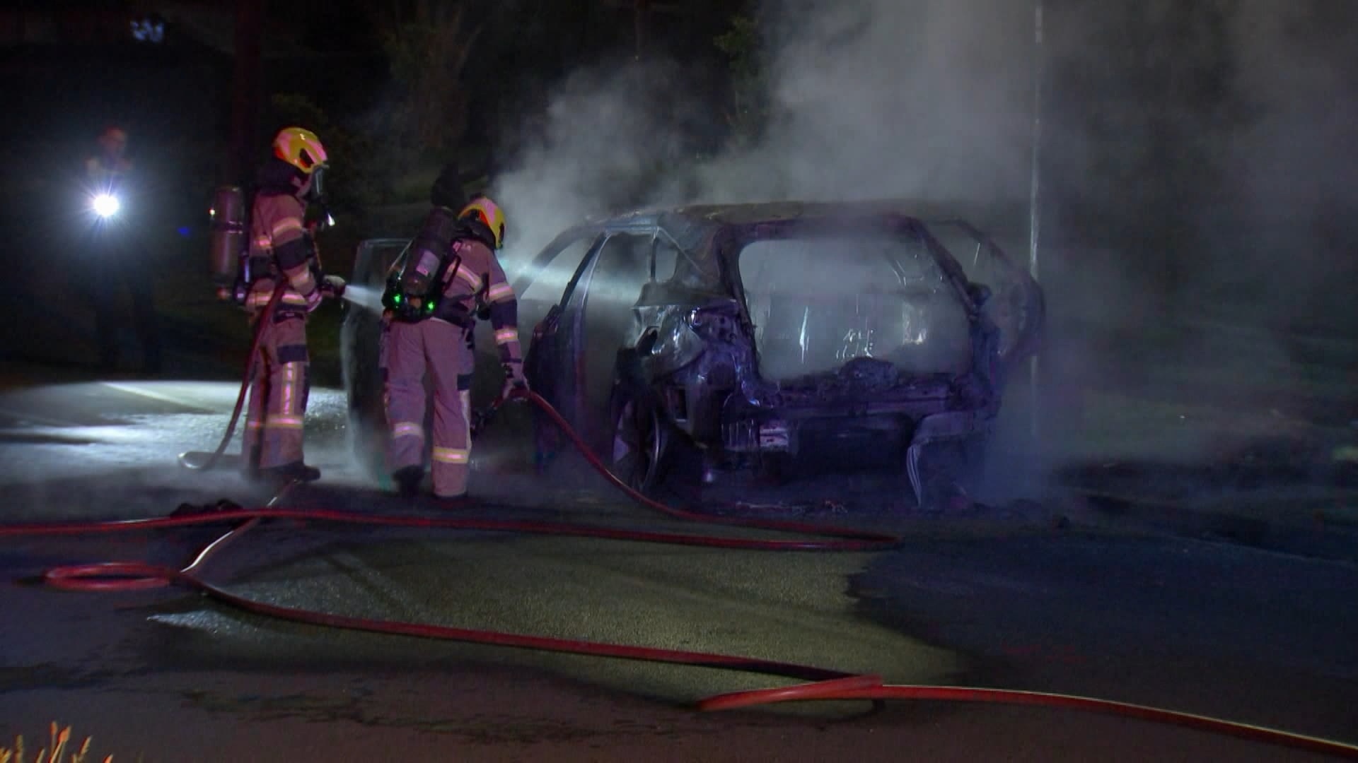 A burnt out car with two firefighters and hoses.