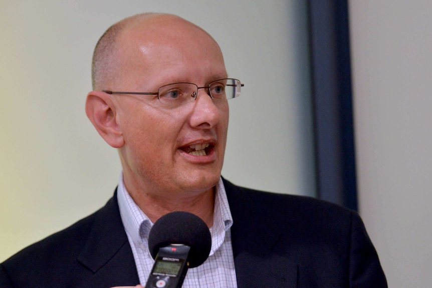 A bald, bespectacled man wearing a suit, apparently speaking into a microphone.
