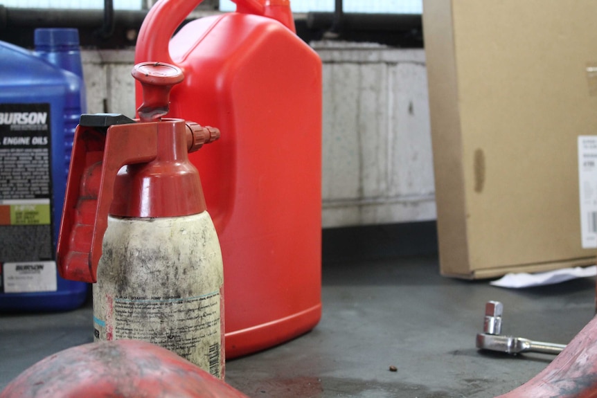 Three bottles, one blue, one white and one red sit on a mechanic shop bench. The white bottle is in focus.