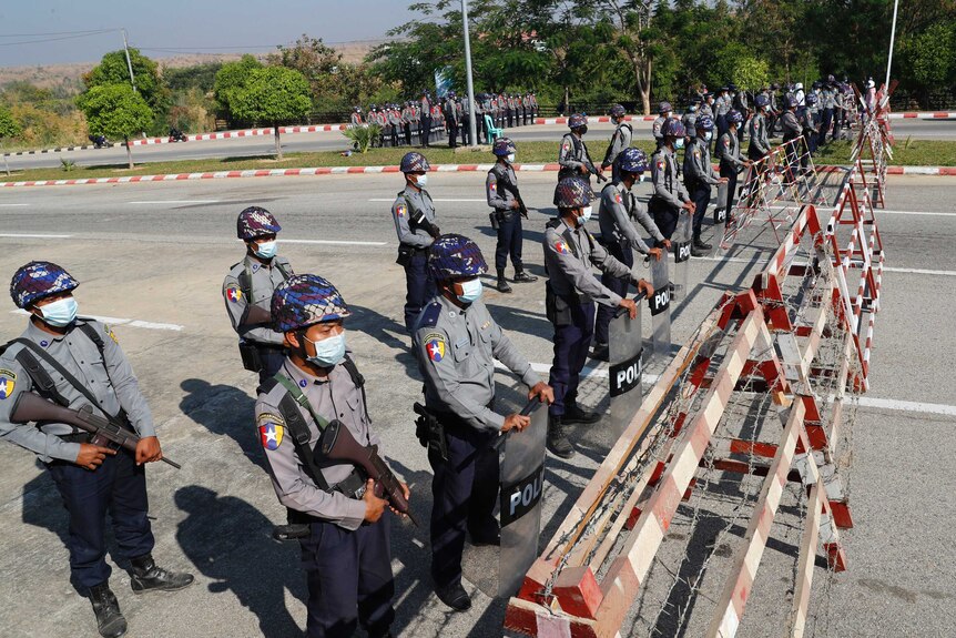 A line of people dressed in police uniforms holding shields and wearing helmets and face masks stand on a road.