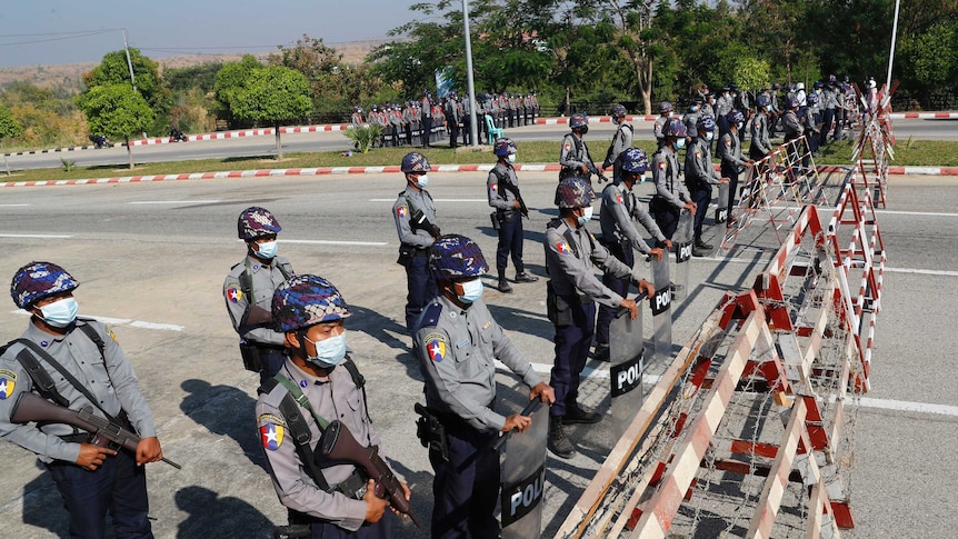 A line of people dressed in police uniforms holding shields and wearing helmets and face masks stand on a road.