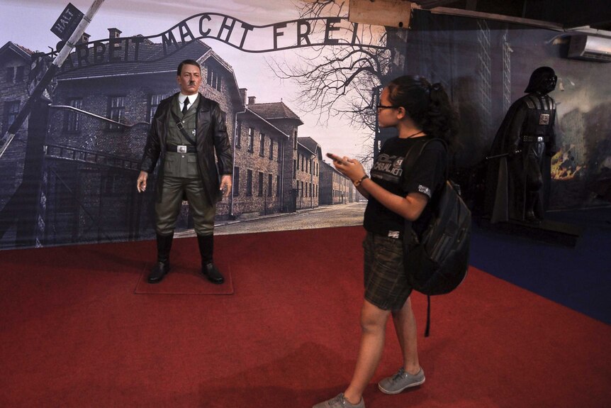 A museum visitor looks at a wax statue of Adolf Hitler standing in front of an image of Auschwitz-Birkenau concentration camp.
