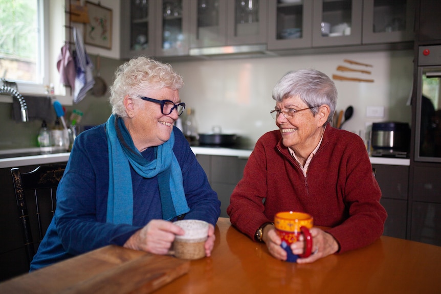 Two women in their 70s holding tea cups laugh while looking at each other.