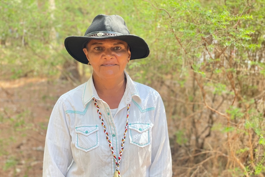 A woman wearing a dark, broad-brimmed hat stands in bushland.