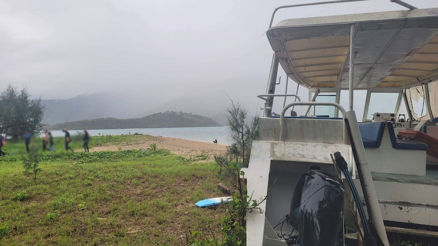 A large boat sits on a grassy beach, window shades are torn and mildew and rust grow on the boat