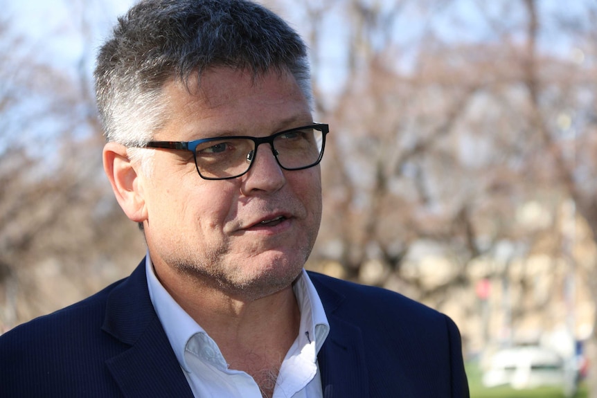 A head and shoulders shot of a grey-haired, bespectacled man in dark suit jacket and a white shirt, open at the throat.