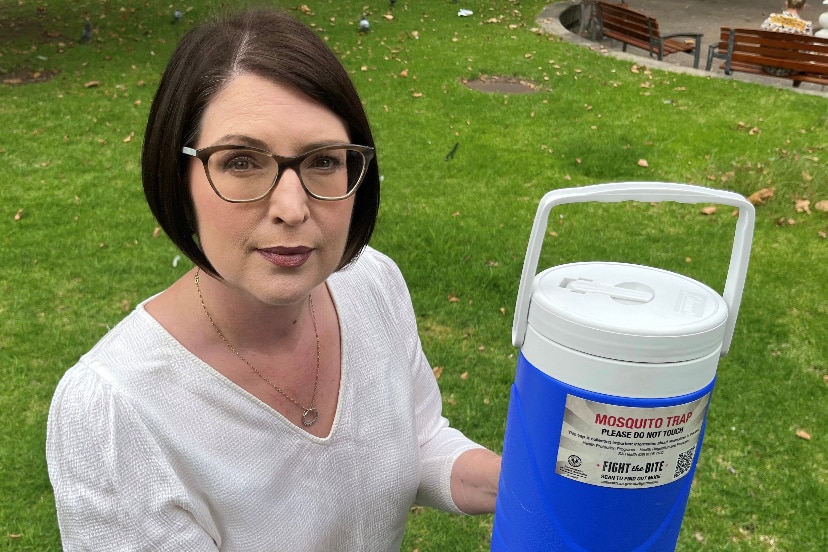 A bespectacled woman with dark hair holding a cylindrical, esky-like vessel that says "mosquito trap" on it.