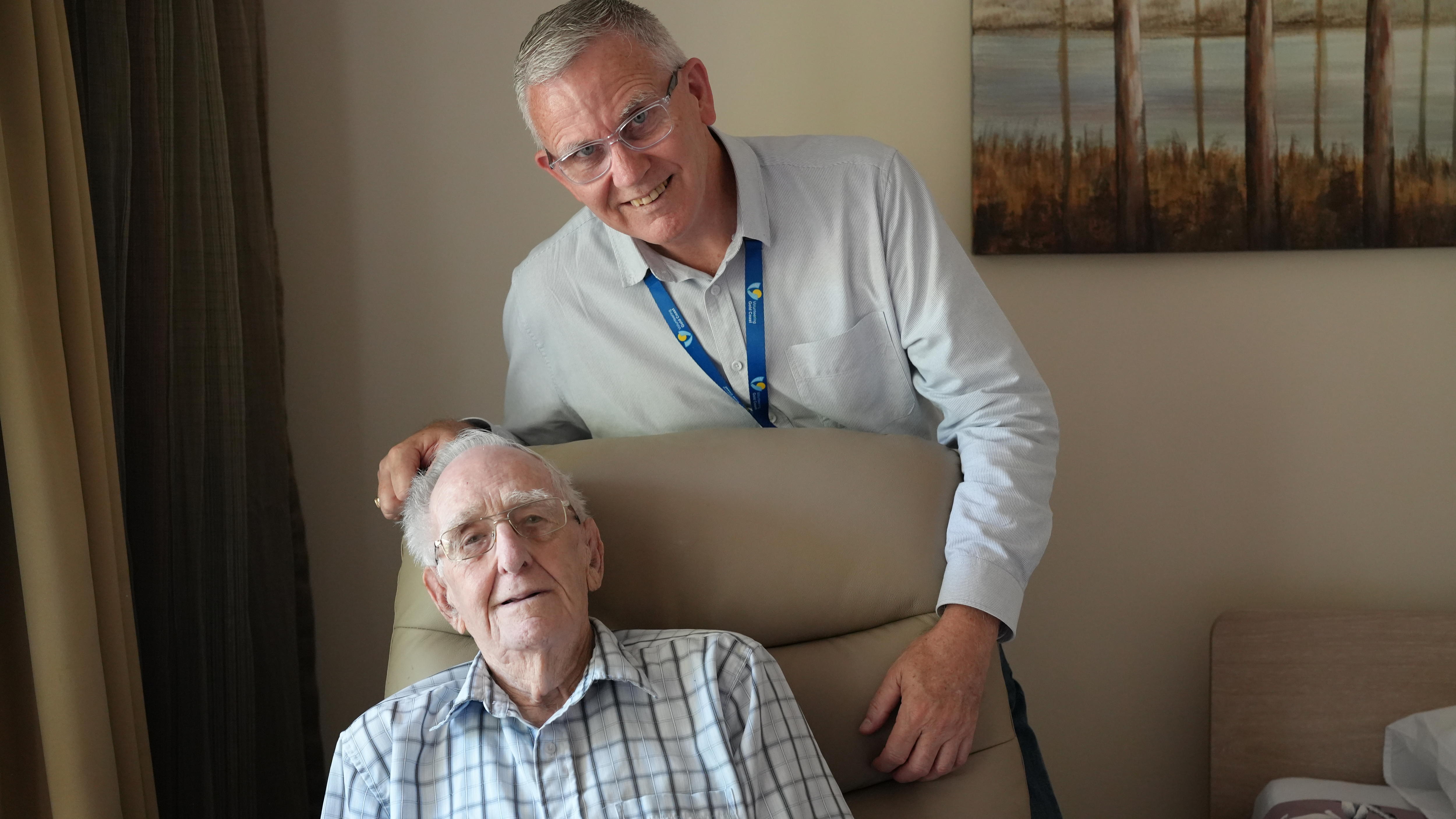 A man leans over the back of another man sitting in a chair.