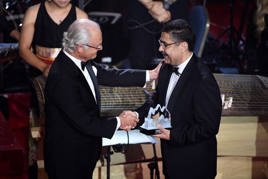 Two men in tuxedos shake hands on a stage, one holding a trophy