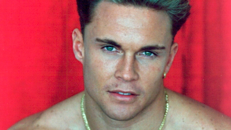 Brett Boyd without a shirt and wearing a gold chain poses for the camera in a modelling glamour shot.