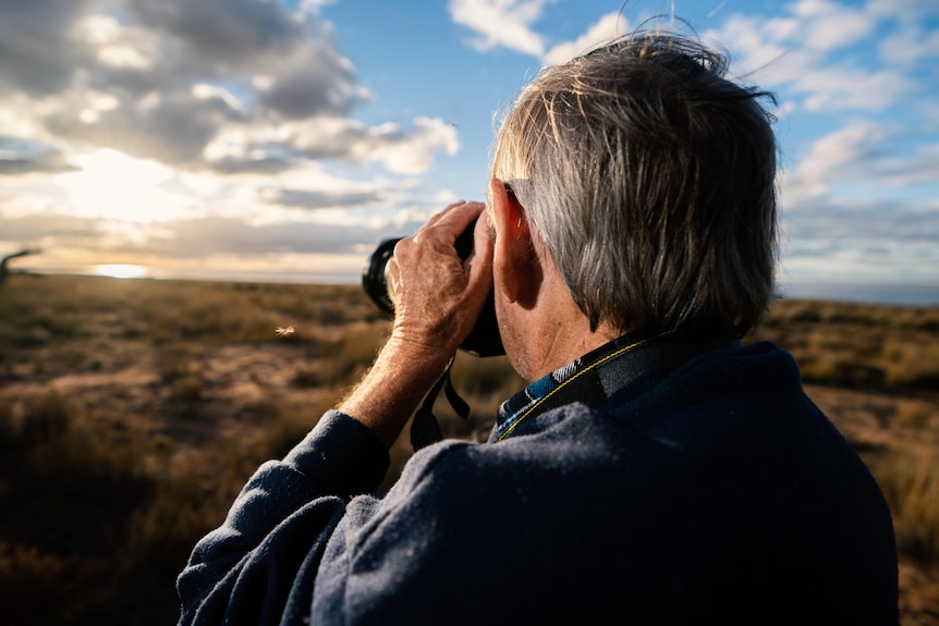 view from behind of man holding a camera to his eyes and shooting across the landscape 