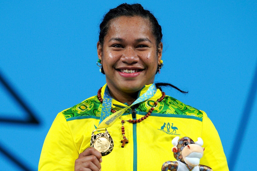 weightlifter eileen cikamatana holds a gold medal and a small mascot toy on top of a commonwealth games podium smiling