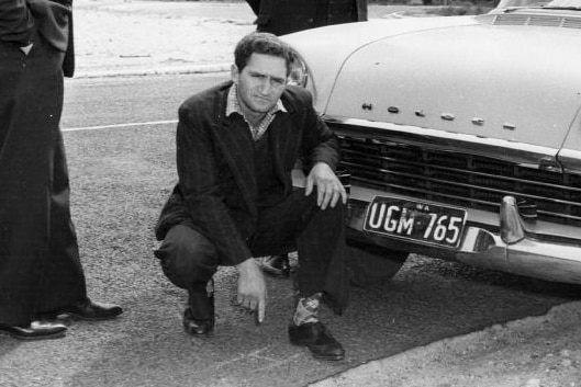 A black and white pic of Eric Edgar Cooke, crouching in front of a police car.