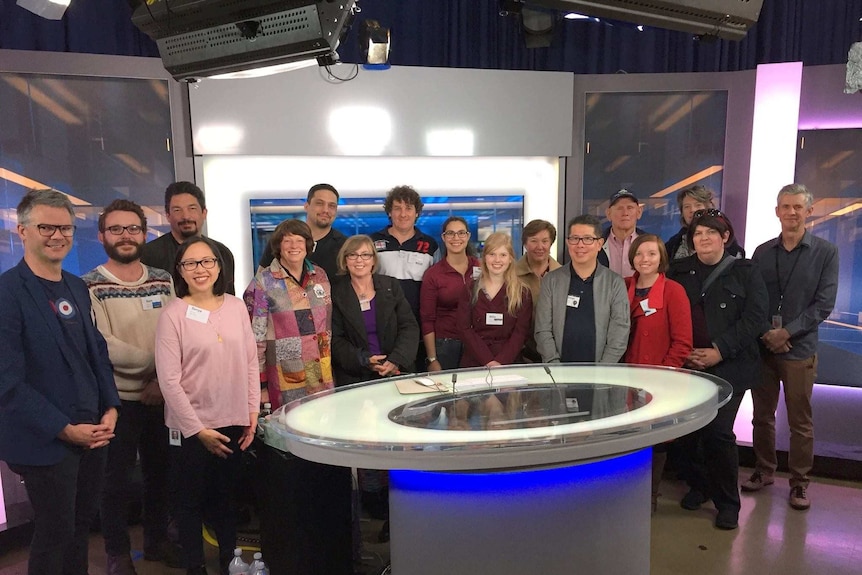 17 people who joined the tour pose for a photo while standing behind the news desk in the ABC studio.