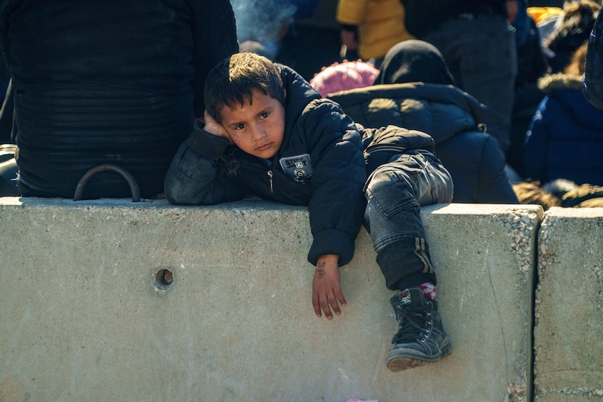 A young boy wearing a black puffer jacket lies on a wall near the Syrian border.